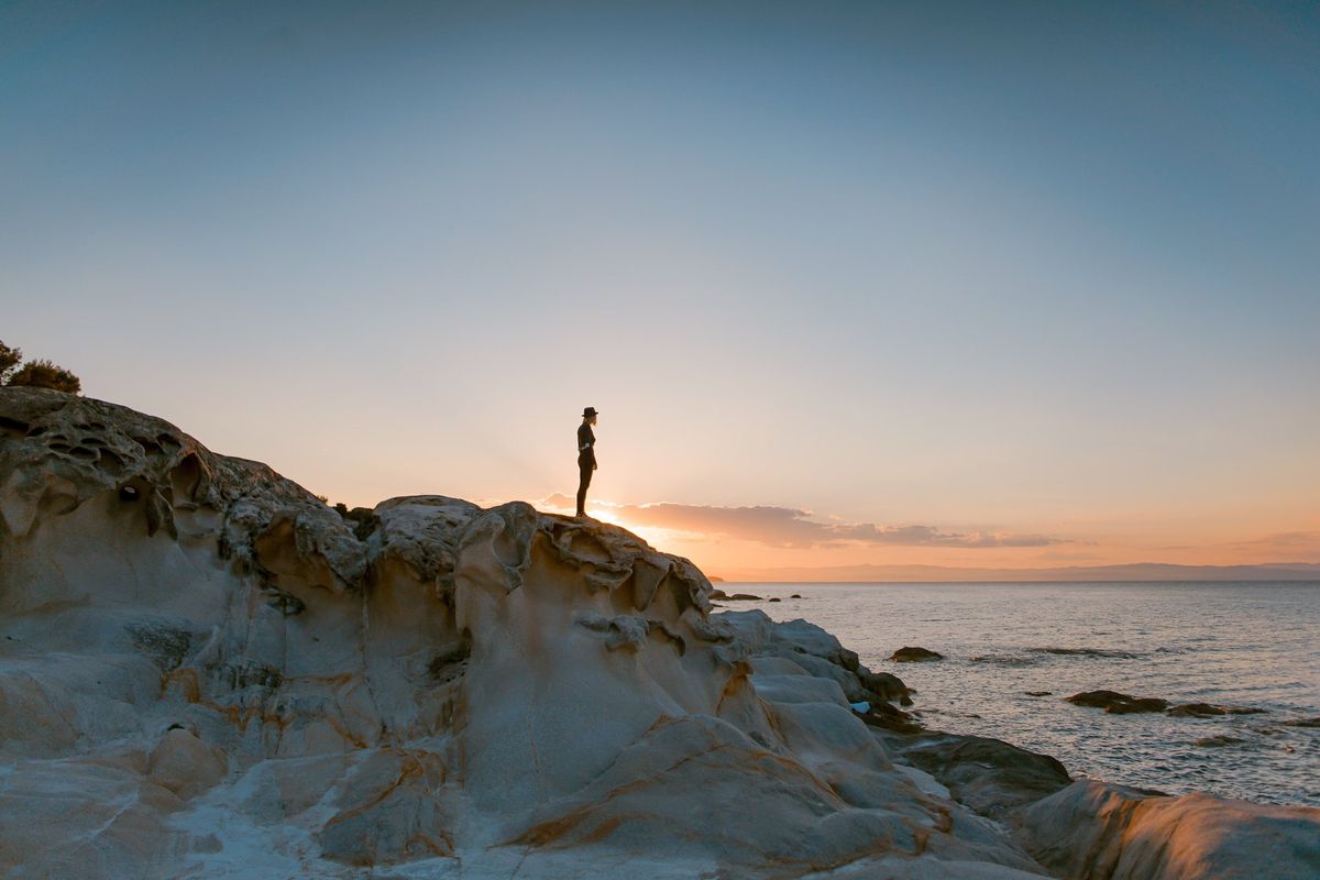 Person standing on a rocky Greek coastline at sunset